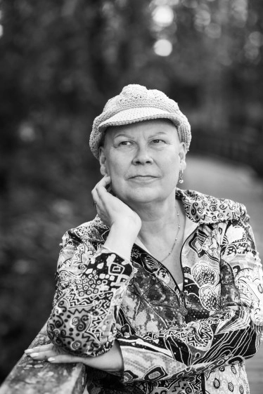 Mustavalkoinen lähikuva Eeva-Kaisasta, jolla on virkattu hattu ja kuviollinen takki, nojaamassa puiseen kaiteeseen ulkona.